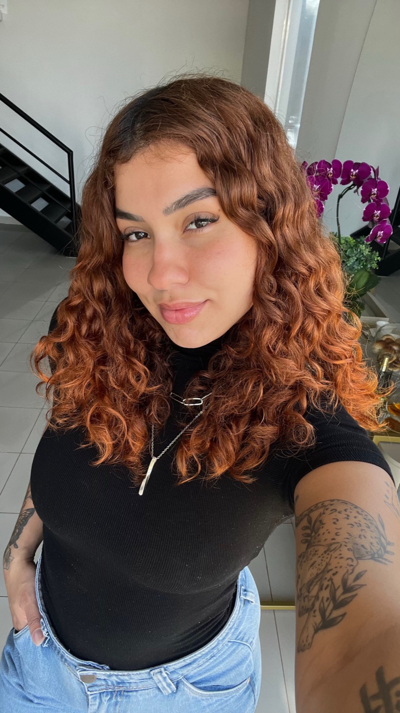 Victoria sorrindo em ambiente claro, cabelo cacheado ruivo.